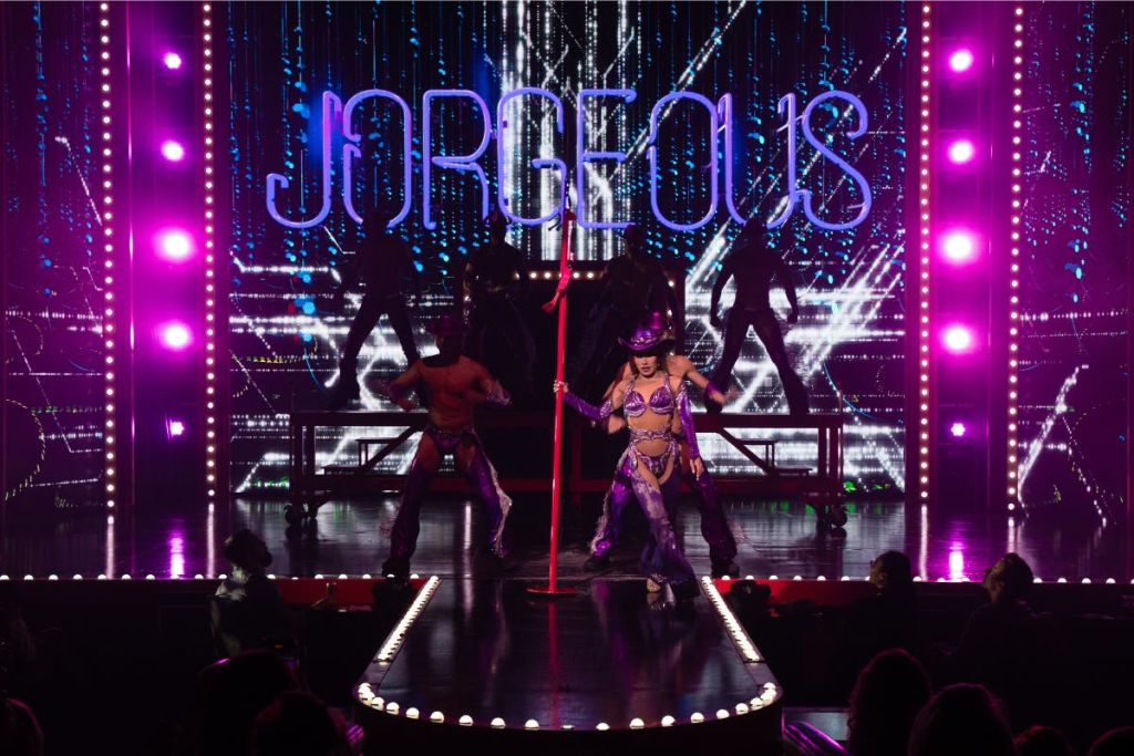 무대 뒤에서 "JORGEIOUS" 가 켜진 보라색 복장과 모자 춤을 추는 연주자, 그림자 속에서 예비 댄서들이 함께합니다.