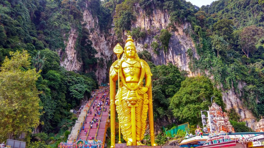 울창한 녹음과 바위 절벽으로 둘러싸인 사람들이 오르는 가파르고 화려한 계단 아래에 신의 황금상이 서 있습니다.