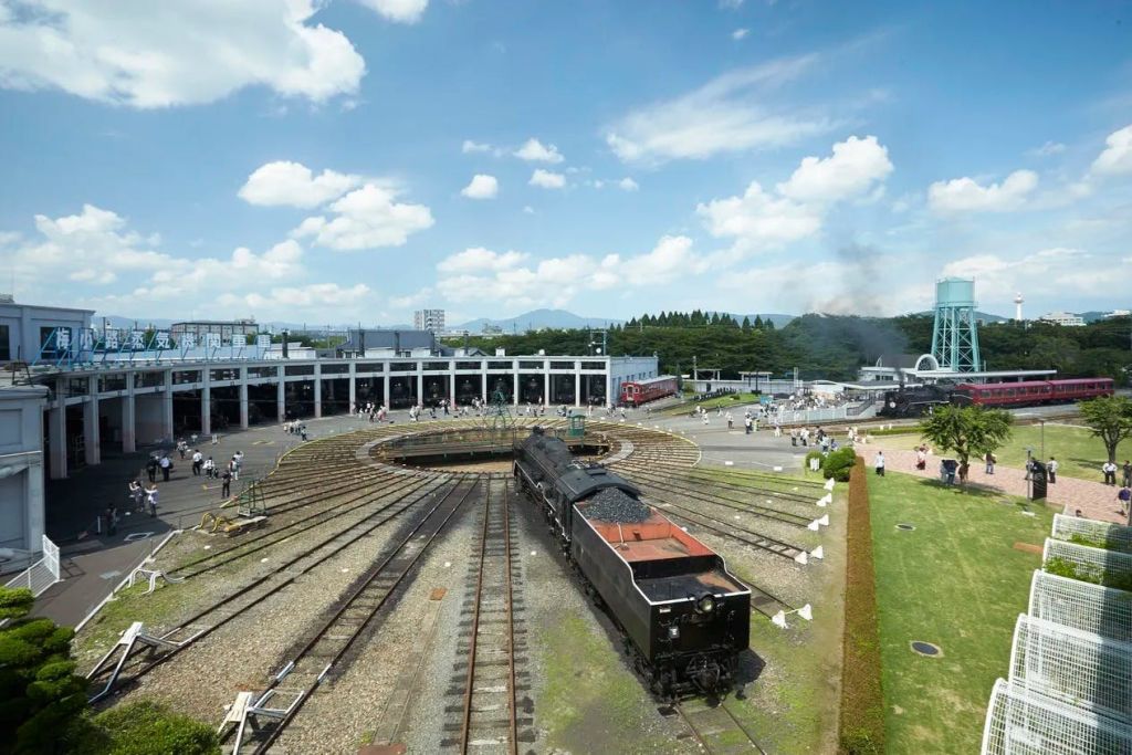 턴테이블과 다양한 철길이 있는 라운드하우스로 검은색 증기 기관차와 근처를 걷는 사람들이 있습니다.