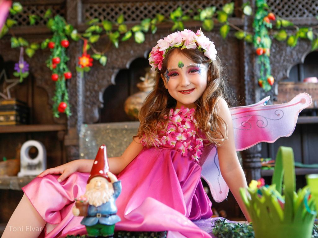 요정 의상과 화관을 쓴 소녀가 활기찬 실내 장식 식물로 둘러싸인 그놈을 들고 미소를 짓고 있습니다.