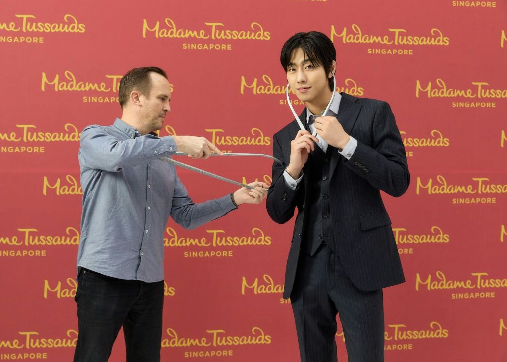 파란색 셔츠를 입은 남성이 'Madame Tussauds Singapore' 브랜딩으로 빨간색 배경에 수트를 입은 왁스 피규어를 측정합니다.