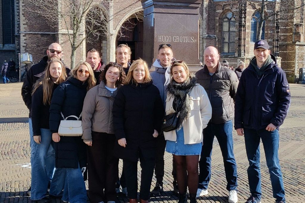 한 무리의 사람들이 "휴고 그로티우스" 라는 글씨가 새겨진 조각상 앞에서 야외 도심에서 포즈를 취하고 있습니다.