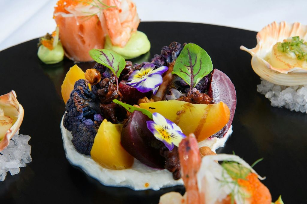 알록달록한 야채와 식용 꽃, 장식품을 검은 접시에 정리한 고급 요리입니다.
