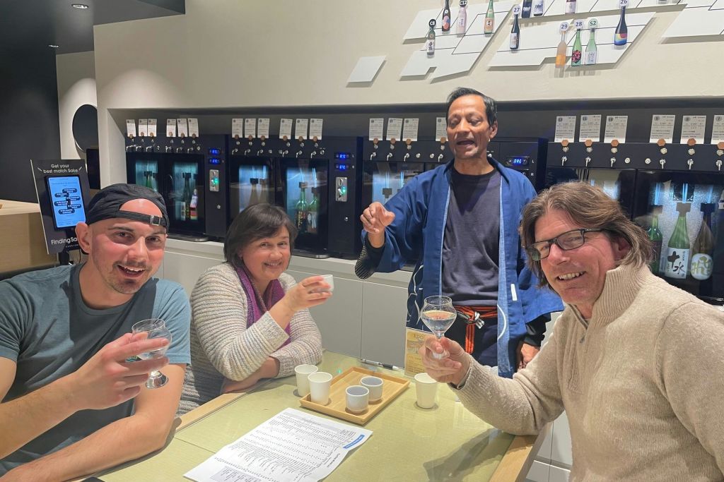 사케 테이스팅이 준비된 테이블에서 잔을 들어 올리는 네 명의 미소 짓는 사람들. 배경은 사케 디스펜서와 디스플레이 병을 보여줍니다.