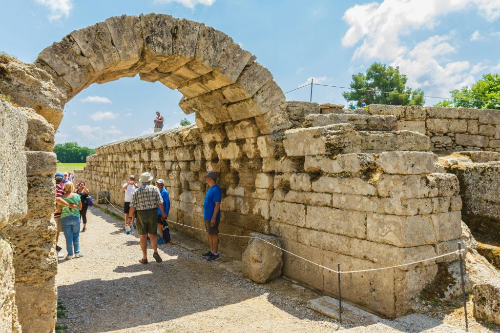 고대 석조 아치형 통로와 벽의 잔해 근처에 서서 맑고 푸른 하늘을 배경으로 한 무리의 사람들.