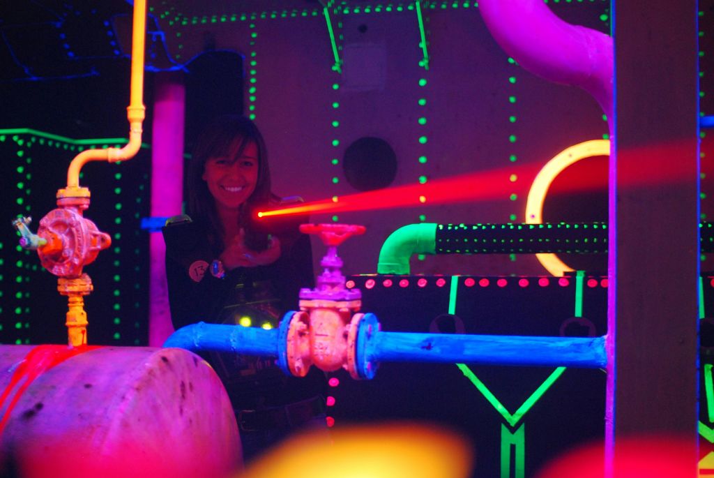 빨간색 레이저 빔을 방출하는 장치를 들고 빛나는 파이프와 밸브가 있는 네온 조명 환경에서 웃고 있는 사람.