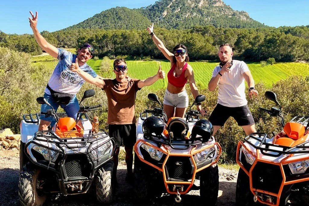 푸른 들판과 숲이 우거진 언덕을 배경으로 오렌지색 ATV 두 대를 들고 포즈를 취하는 네 사람.