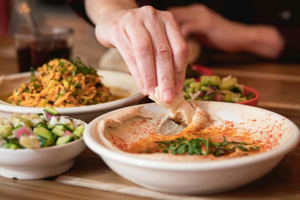 나무 테이블 위에 샐러드와 야채 접시가 놓인 후무스 그릇에 빵을 담그는 손.