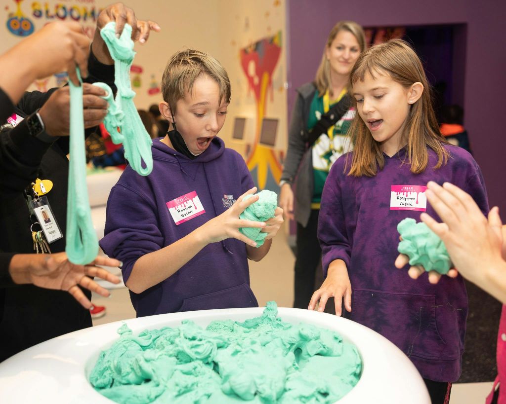 보라색 후드를 입은 두 명의 어린이가 식탁에서 성형이 가능한 녹색 거품을 가지고 놀고 있으며, 이를 배경으로 어른이 감독합니다.