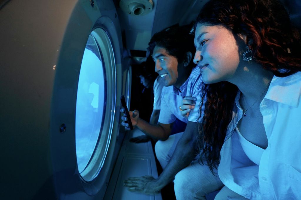 잠수함의 두 사람이 수중 장면에서 둥근 창문을 내다보고 있습니다. 한 사람은 휴대폰을 들고 미소를 짓고 있습니다. 파란색 조명이 공간을 가득 채웁니다.