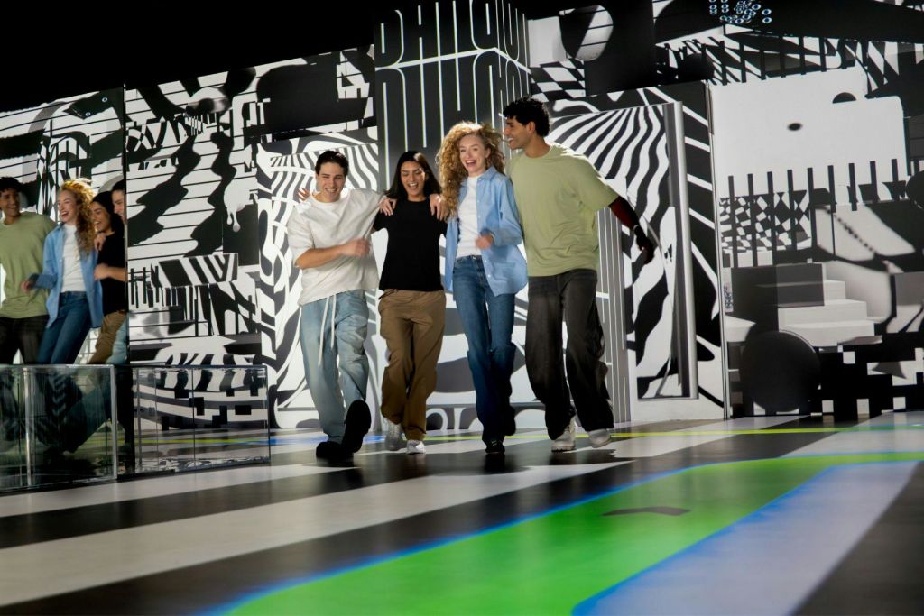 흑백 디자인과 화려한 바닥 액센트가 돋보이는 활기찬 패턴의 환경에서 네 명이 함께 걷고 있습니다.