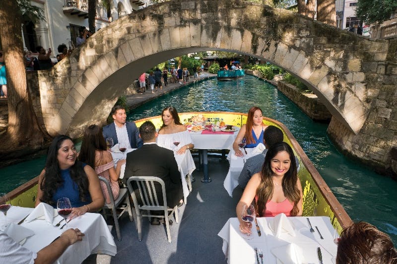햇살이 내리쬐는 야외 환경에서 녹지로 둘러싸인 운하의 돌다리 아래에서 보트를 타고 식사를 즐기는 사람들.
