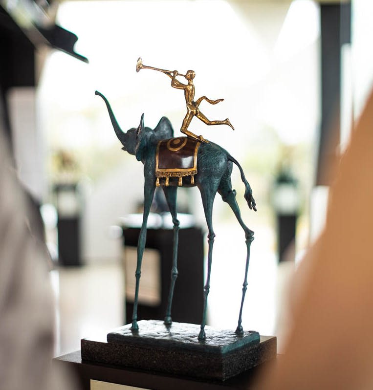 얇고 긴 다리의 코끼리가 안장을 짚고 트럼펫을 연주하는 모습을 담은 청동 조각상.