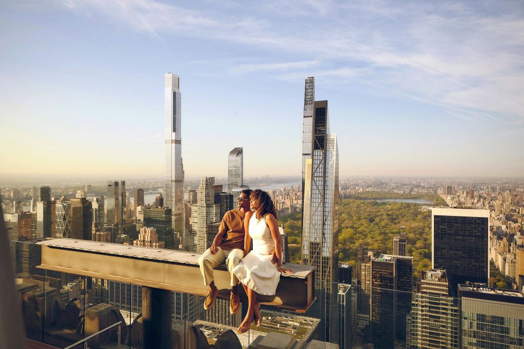 높은 건물이 있는 도시 풍경과 푸른 하늘 아래 멀리 떨어진 공원이 내려다보이는 고층빌딩 꼭대기의 대들보에 한 커플이 앉아 있습니다.
