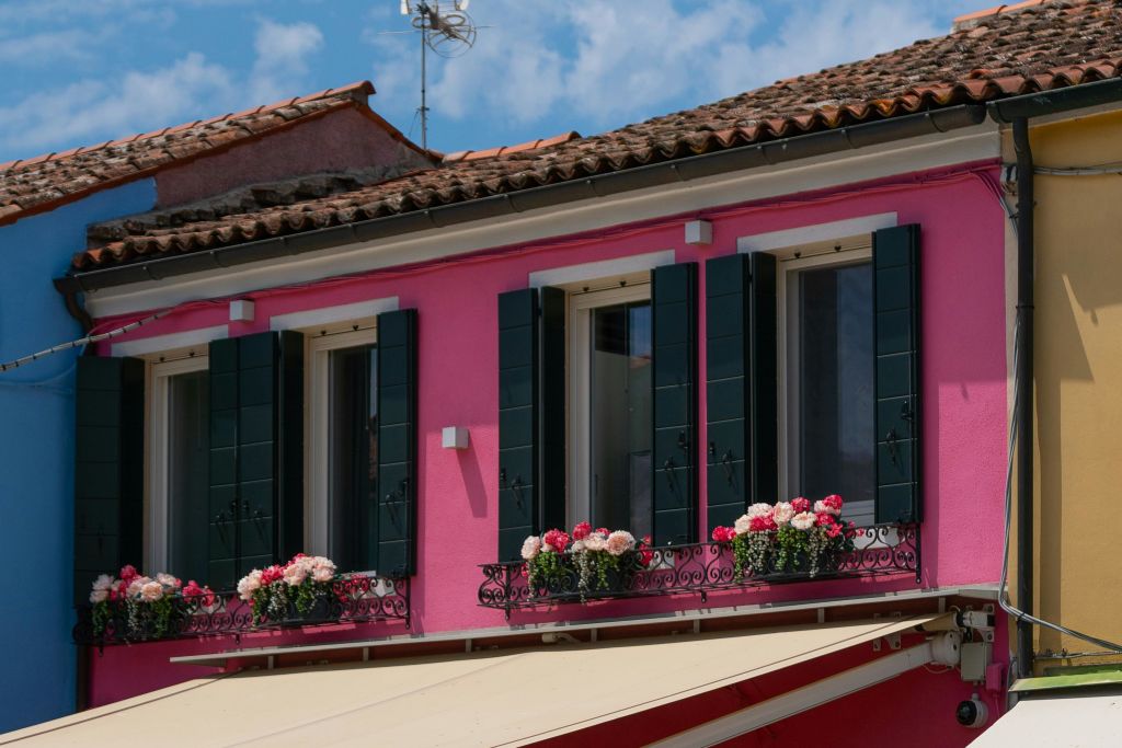 타일로 덮인 지붕 아래 상층 창문 3개, 초록색 셔터, 분홍색과 흰색 꽃이 가득한 꽃 상자가 있는 분홍색 주택입니다.