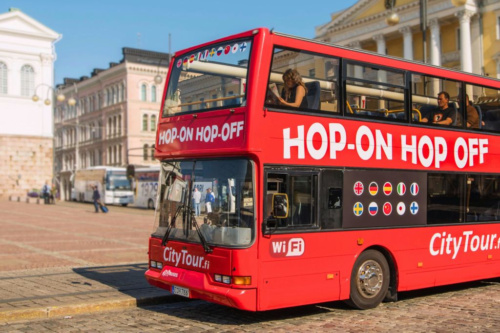 측면에 "홉온 홉오프 (hop-on hop-off)" 라고 쓰여진 도시 광장의 밝은 빨간색 2층 투어 버스. 분주한 도시 배경.