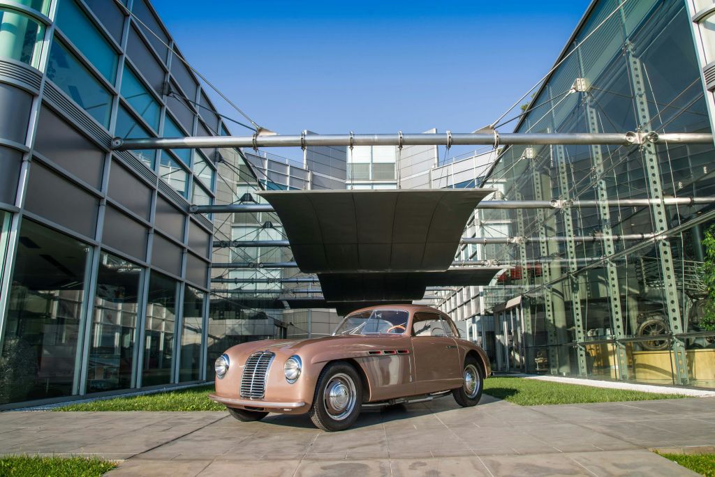 화창한 날 검은색 대형 캐노피 아래 현대적인 유리 건물 앞에 빈티지 브라운 차량이 주차되어 있습니다.