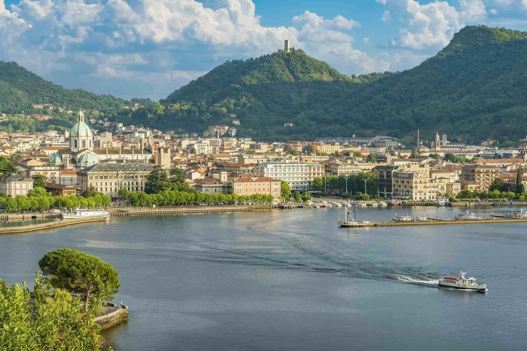 건물, 언덕 꼭대기 타워, 부분적으로 흐린 하늘 아래 푸른 언덕이 있는 호숫가 마을의 경치.