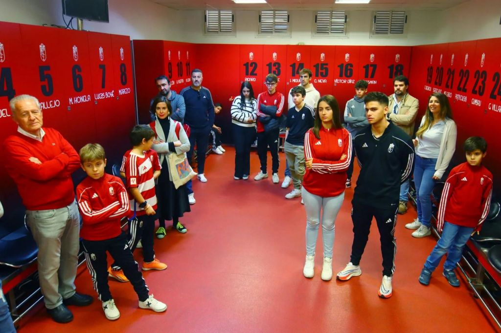 한 무리의 사람들이 번호가 매겨진 사물함이 있는 빨간색 방에 서 있으며, 모두 캐주얼 스포츠웨어를 입고 있습니다. 두 사람이 전경에 중심을 두고 있습니다.