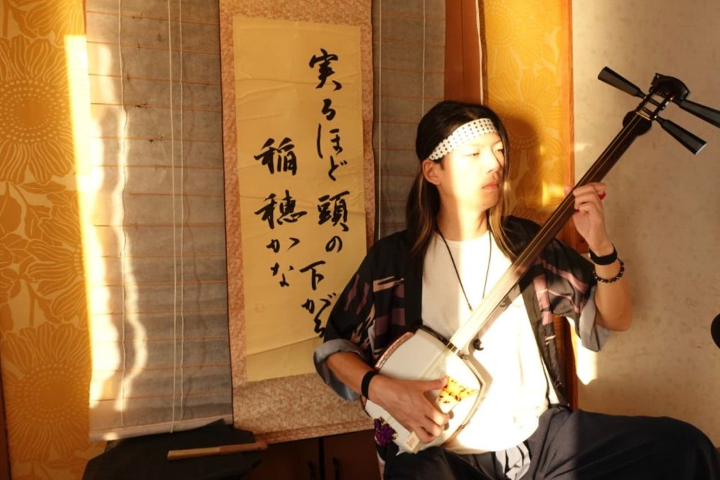 전통 복장을 한 사람이 샤미센을 입고 서예 두루마리를 배경으로 연주하고 있습니다. 빛이 얼굴에 그림자를 드리웁니다.