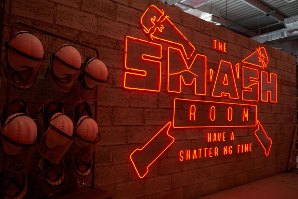 벽돌 벽에 걸린 선반에 안전 헬멧이 달린 "The Smash Room" 이라는 네온 사인. 텍스트에 "산산조각나는 시간 보내기" 가 포함되어 있습니다.