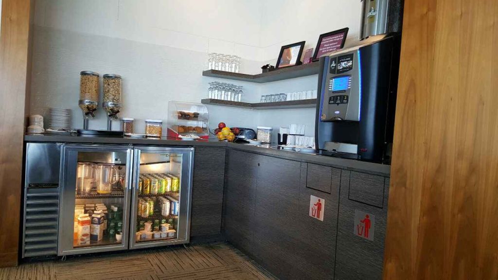 커피 머신, 음료 냉장고, 과일, 페이스트리, 유리잔과 냅킨이 있는 선반이 있는 휴게실.