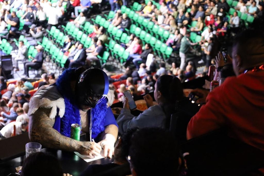 녹색 좌석이 있는 혼잡한 경기장에서 파란색과 검은색 의상을 입은 사람이 사인을 하고 있습니다.