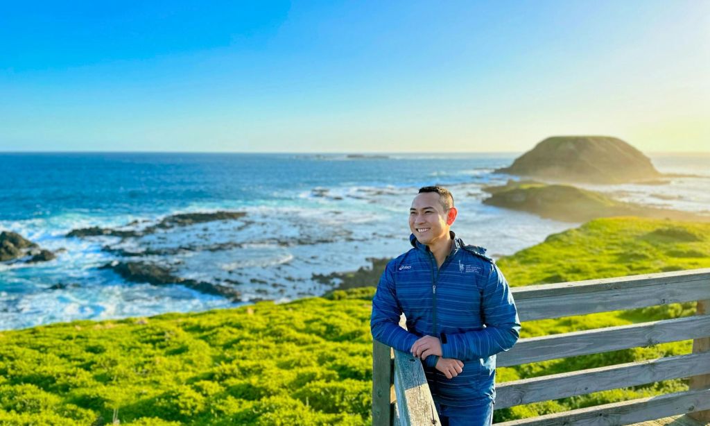 파란색 재킷을 입고 나무 난간에 기대어 웃고 있는 사람으로, 아름다운 해안 풍경을 배경으로 합니다.