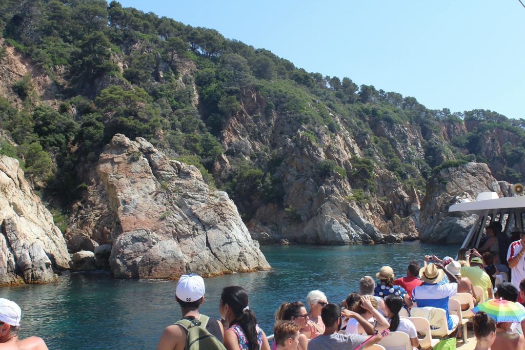 울창한 푸른 나무와 밝은 하늘 아래 맑고 푸른 물이 흐르는 바위 해안 근처에서 보트를 타고 관광하는 관광객.