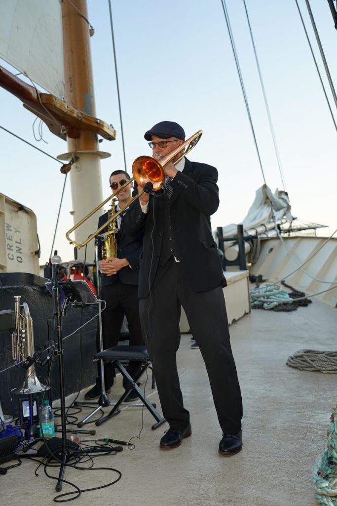 항해하는 배의 갑판에서 돛과 밧줄을 배경으로 금관 악기를 연주하는 두 명의 음악가.