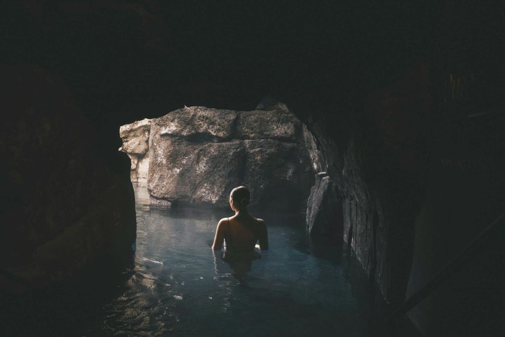 희미한 불빛이 내리쬐는 동굴에서 햇빛이 바위 벽을 비추는 개구부를 바라보며 허리 깊이의 물에 서 있는 사람.