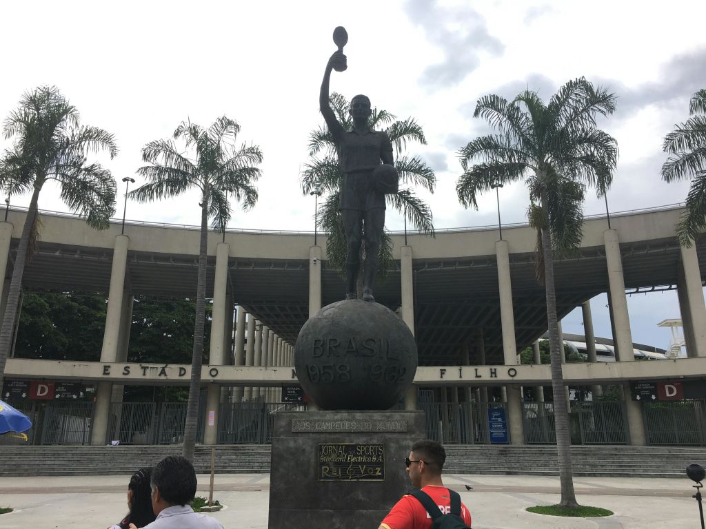 트로피를 들고 있는 축구 선수의 동상으로, 야자수와 전경에 몇 명이 있는 경기장에 세워져 있습니다.