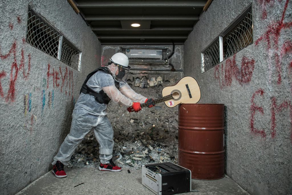 좁고 잔해가 가득한 골목길에서 금속 통에 기타를 부수는 보호 장비를 착용한 사람. 바닥에 고장난 장비.