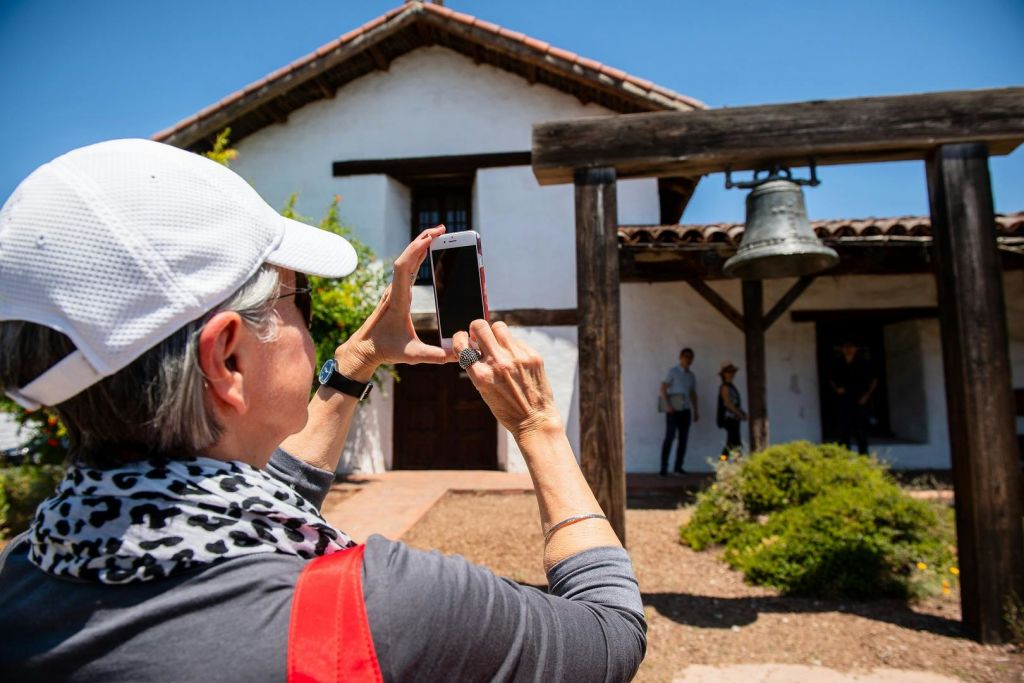 흰색 모자를 쓴 사람이 외부의 커다란 종소리가 있는 역사적인 미션 스타일의 건물을 스마트폰으로 촬영합니다.