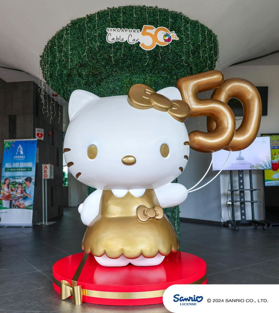 위의 "싱가포르 케이블카 50" 표지판이 있는 빨간색 플랫폼에 표시된 금색 "50" 풍선을 들고 있는 금색 드레스를 입은 헬로 키티 동상.