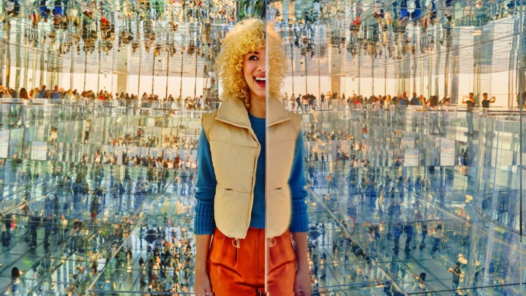 한 사람이 거울 벽 앞에 서서 스플릿 효과를 냅니다. 배경은 사람들의 성찰로 가득 찬 방을 보여줍니다.