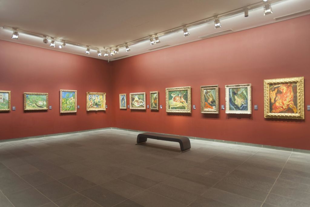 붉은 벽이 있는 박물관 갤러리에 액자 그림이 전시되어 있습니다. 방 중앙의 타일 바닥에 하나의 벤치가 놓여 있습니다.
