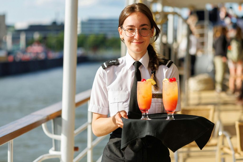 햇살이 내리쬐는 야외 데크에 다채로운 칵테일 두 잔이 놓인 트레이를 들고 있는 유니폼 차림의 미소 짓는 서버.