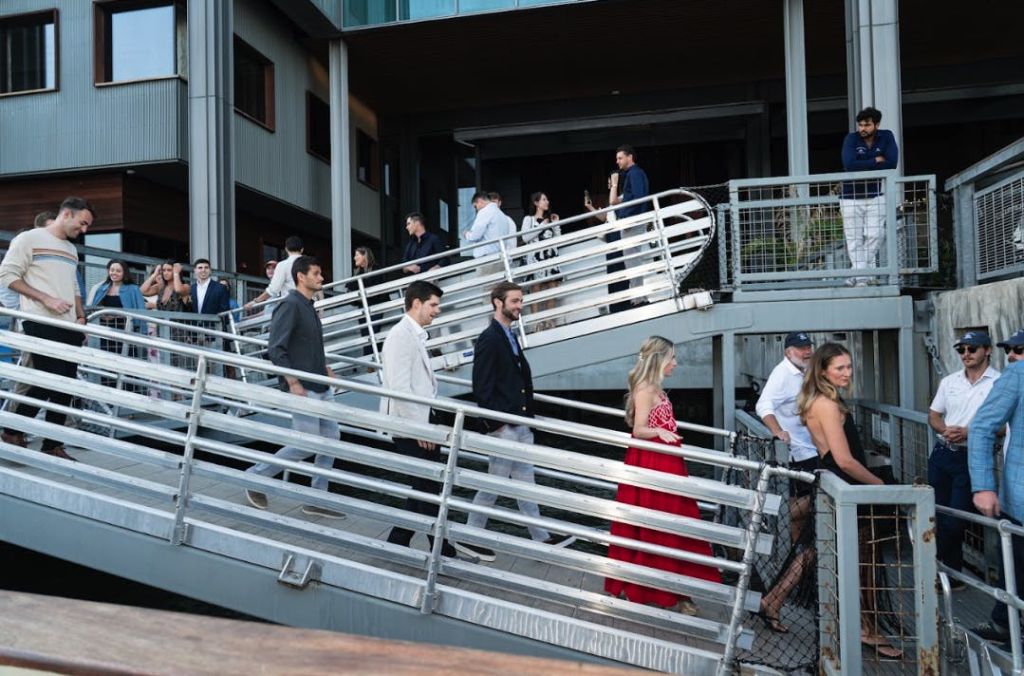 정장 차림의 사람들은 건물 근처의 금속 경사로를 걷고, 다른 사람들은 위층에 서서 대화를 나눕니다.