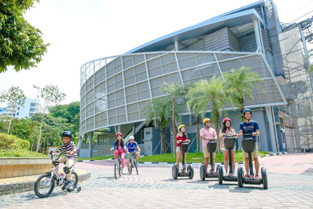 야자수와 타일로 마감된 산책로가 있는 현대적인 건물 앞에서 세그웨이를 타고 자전거를 타는 사람들의 일행.