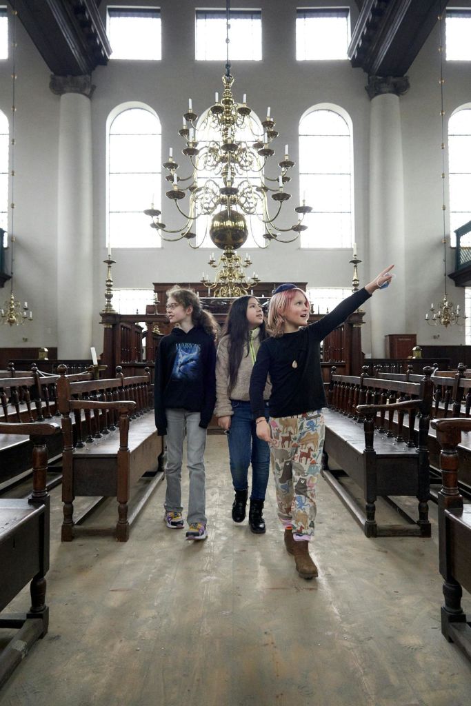 세 명의 어린이가 높은 창문과 화려한 샹들리에가 있는 그랜드 홀의 통로를 따라 걸어갑니다. 한 명의 어린이가 앞을 가리킵니다.