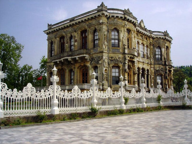 정교한 디자인, 커다란 창문, 장식용 흰색 울타리가 있는 화려하고 유서 깊은 건물입니다. 포장된 공간과 녹지가 전면에 있습니다.