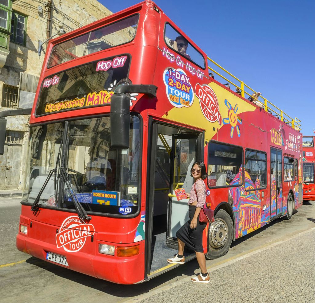 "Official Tour" 와 "Hop On Hop Off" 로고가 있는 빨간색 2층 투어 버스가 선글라스를 쓴 사람이 주차되어 있습니다.