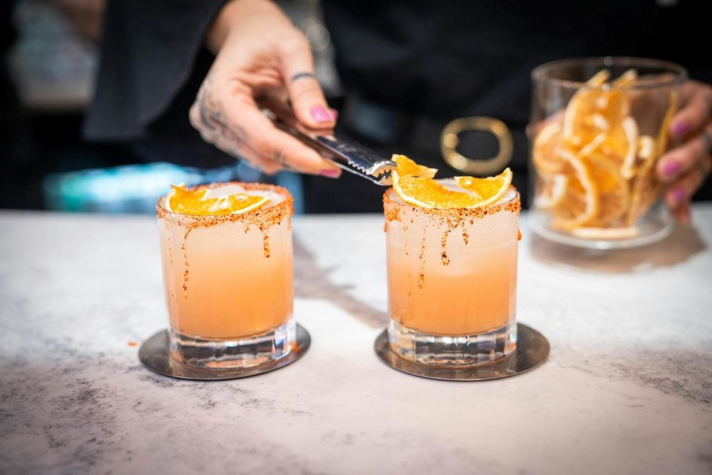 오렌지 슬라이스와 설탕 테두리가 있는 두 개의 칵테일을 손으로 집게로 장식합니다. 배경에 주황색 조각이 담긴 항아리가 흐릿하게 보입니다.