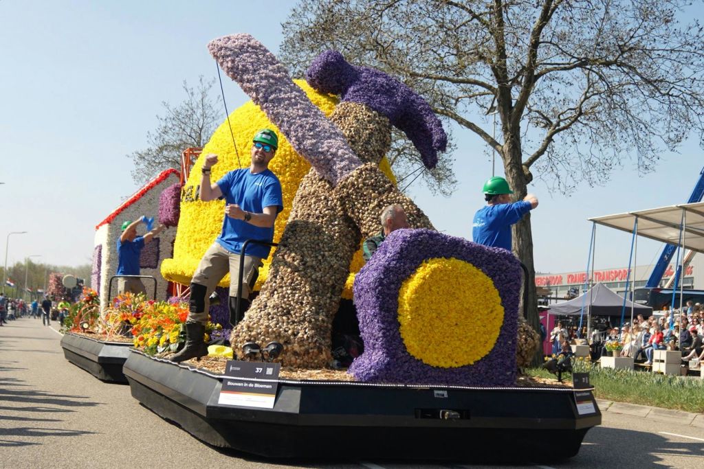 파란색 셔츠와 녹색 헬멧을 착용한 남성은 망치와 건축 테마를 형상화한 커다란 꽃으로 장식된 수레를 타고 있습니다.