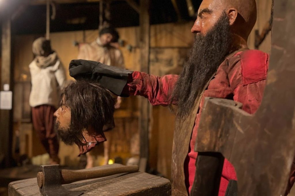 빨간색 옷을 입은 마네킹으로 긴 수염에 소품 머리를 들고 있으며, 나무 장식이 있는 역사적이거나 테마가 있는 전시품의 일부입니다.