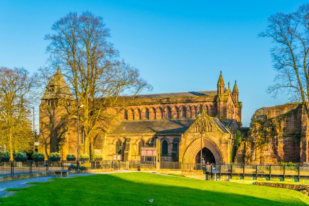아치형 입구, 커다란 창문, 탑이 있는 역사적인 석조 건물로, 맑고 푸른 하늘을 배경으로 잔디가 우거진 지역과 경계를 이루고 있습니다.