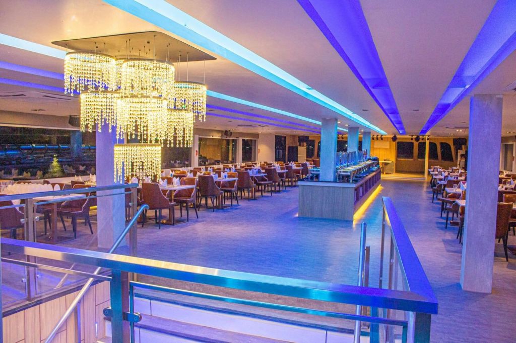 푸른 조명, 샹들리에, 깔끔하게 정리된 테이블, 뷔페 카운터가 있는 현대적인 레스토랑입니다. 전면에 유리 난간이 있는 계단.