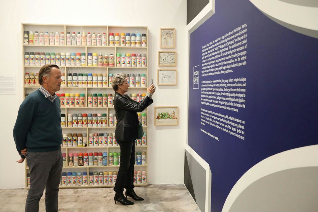 벽면 사진을 찍는 여성과 스프레이 페인트 캔이 가득한 선반 근처에 서 있는 남성, 두 사람이 갤러리에 있습니다.