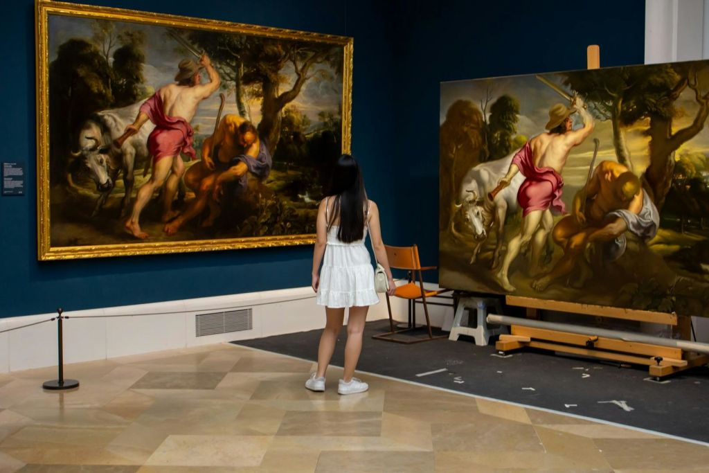 흰색 드레스를 입은 한 여성이 파란색 벽이 있는 미술관에 전시된 두 개의 커다란 고전 그림을 관찰합니다.